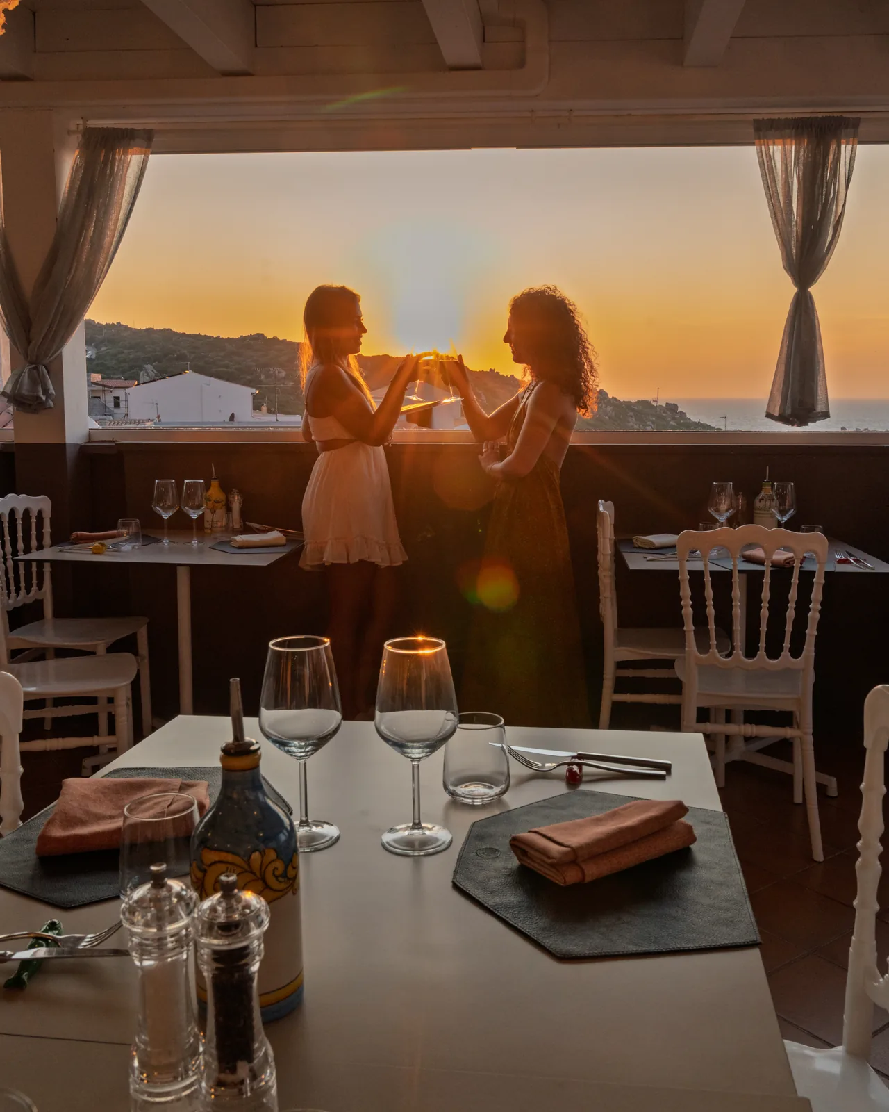 Tavolo apparecchiato in terrazza al tramonto, con due ospiti che brindano sul fondo